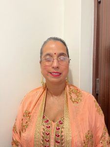 Amrit is a middle aged south Asian woman who is smiling towards the camera against a white background.  She is wearing an Indian outfit which you can see the top half, a peach and gold top with red in the boarder.  She is wearing matching gold earrings, a red bindi and red lipstick.