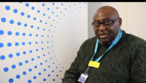 Dr Sylvanus’s image.: Dr Sylvanus, a black middle aged man who is bold and is wearing glasses, is smiling and looking towards the camera.  He is wearing a dark green wollen jumper with a lanyard. He is on the right and to the left of him, you can see a white background with blue dots.