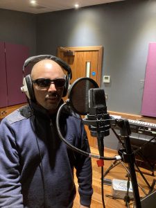 Talat is standing in front of a microphone. He is wearing a navy blue top and headphones with black sunglasses. You can see a door behind him.