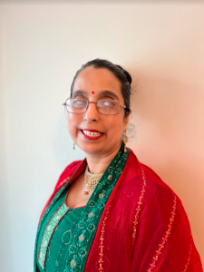 Headshot image of Amrit smiling. Wearing a jade green and bright red traditional suit which is heavily embroidered with gold thread and beading. She is wearing a gold necklace and earrings to match.